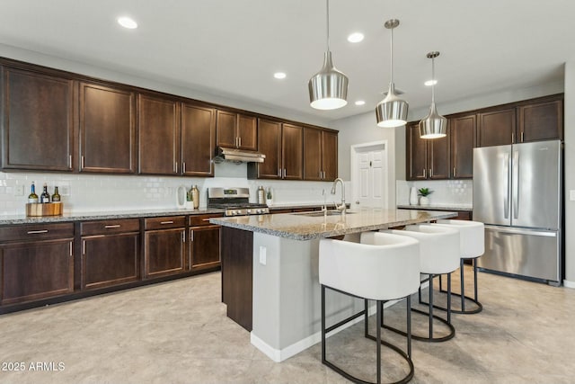 kitchen with sink, hanging light fixtures, a kitchen island with sink, stainless steel appliances, and light stone countertops