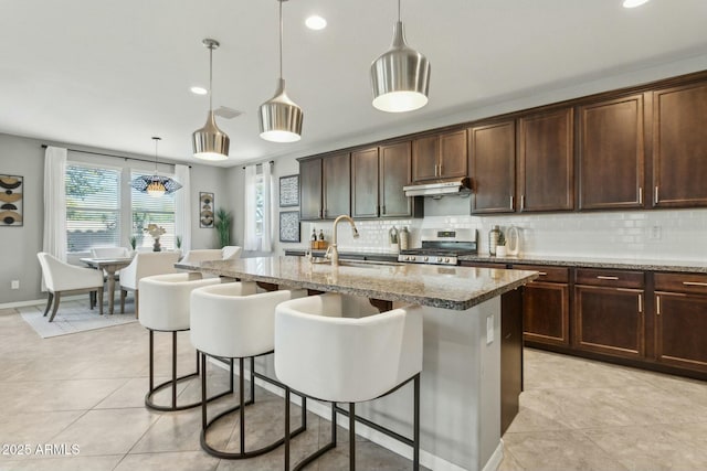 kitchen featuring sink, a center island with sink, stainless steel range, pendant lighting, and light stone countertops