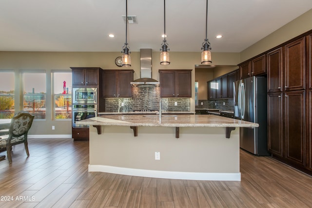 kitchen with pendant lighting, wall chimney exhaust hood, appliances with stainless steel finishes, and an island with sink