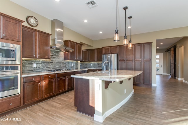 kitchen with an island with sink, appliances with stainless steel finishes, a kitchen bar, light hardwood / wood-style floors, and wall chimney exhaust hood