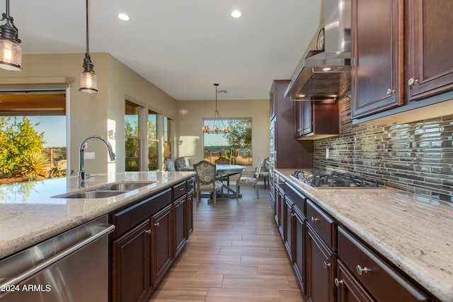 kitchen featuring wall chimney range hood, hanging light fixtures, stainless steel appliances, sink, and light stone counters