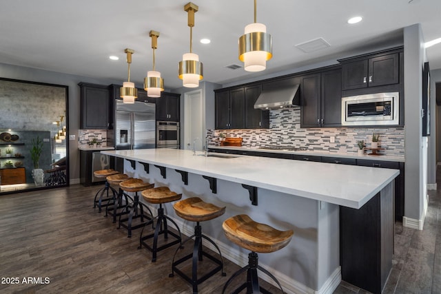 kitchen with decorative light fixtures, sink, built in appliances, a center island with sink, and wall chimney exhaust hood