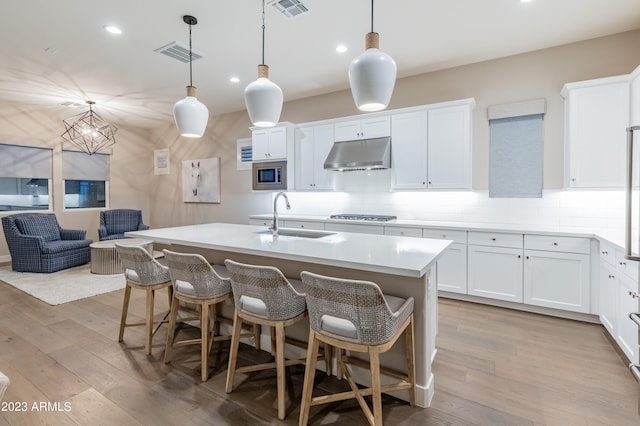 kitchen featuring a kitchen island with sink, white cabinets, sink, light wood-type flooring, and decorative light fixtures