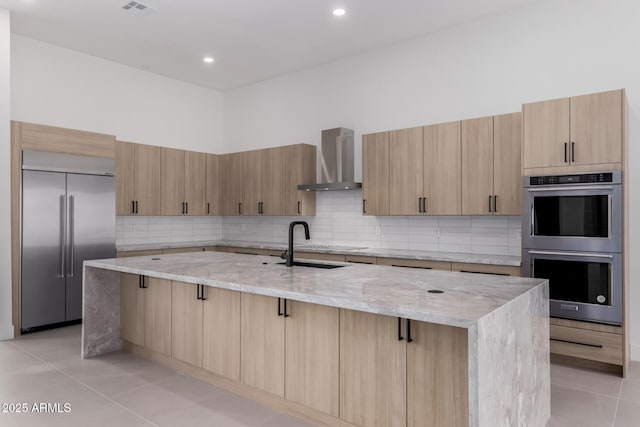 kitchen featuring light stone counters, stainless steel appliances, an island with sink, and wall chimney range hood