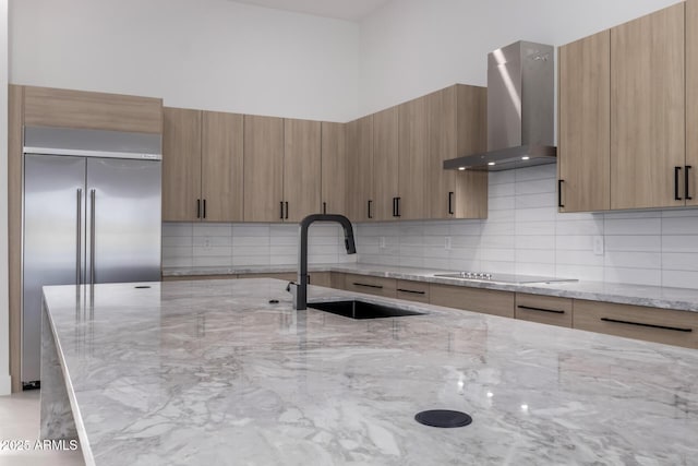 kitchen with built in refrigerator, sink, wall chimney range hood, and light stone counters