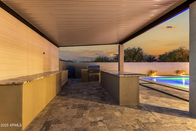 patio terrace at dusk with an outdoor kitchen, a fenced in pool, and area for grilling