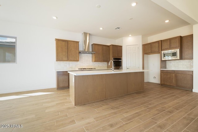 kitchen with stainless steel appliances, wall chimney exhaust hood, brown cabinetry, and light countertops