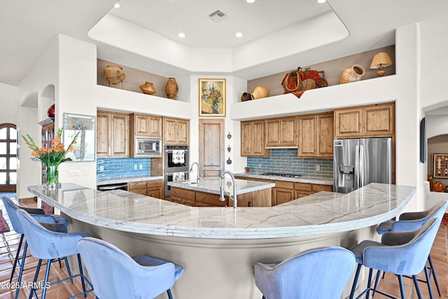 kitchen featuring arched walkways, appliances with stainless steel finishes, a large island with sink, and brown cabinetry