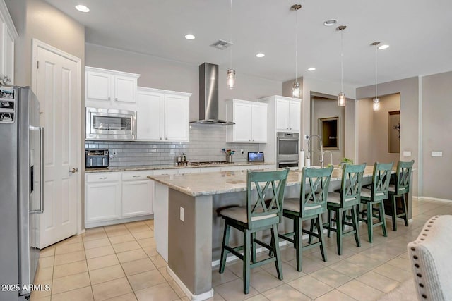 kitchen featuring decorative light fixtures, wall chimney range hood, stainless steel appliances, and an island with sink