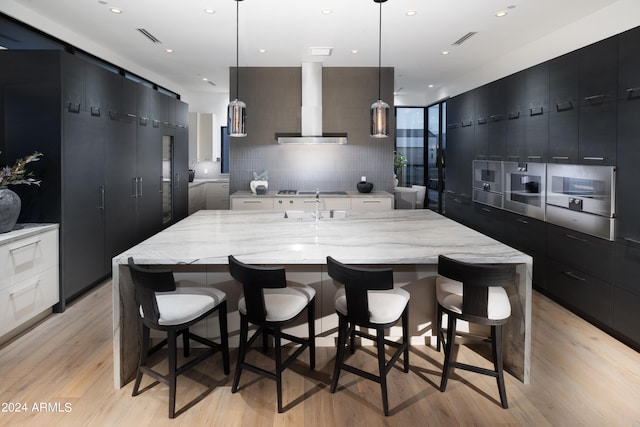 kitchen with light wood-type flooring, tasteful backsplash, hanging light fixtures, wall chimney range hood, and a spacious island