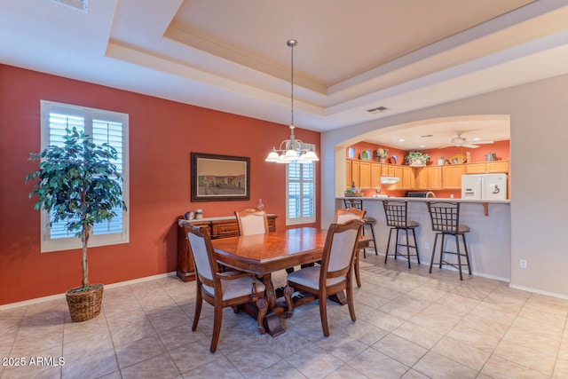 tiled dining area with ceiling fan with notable chandelier, a wealth of natural light, and a tray ceiling