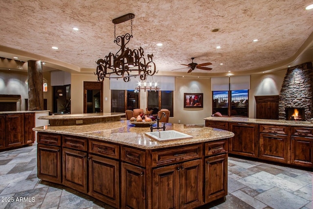 kitchen with pendant lighting, sink, light stone counters, dark brown cabinets, and a center island with sink