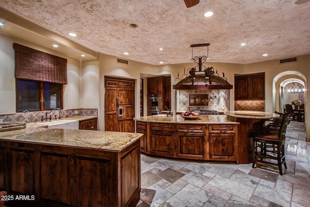 kitchen with sink, light stone counters, hanging light fixtures, a large island with sink, and backsplash