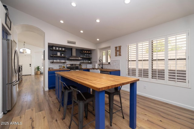 kitchen featuring wood counters, blue cabinets, lofted ceiling, stainless steel appliances, and wall chimney exhaust hood