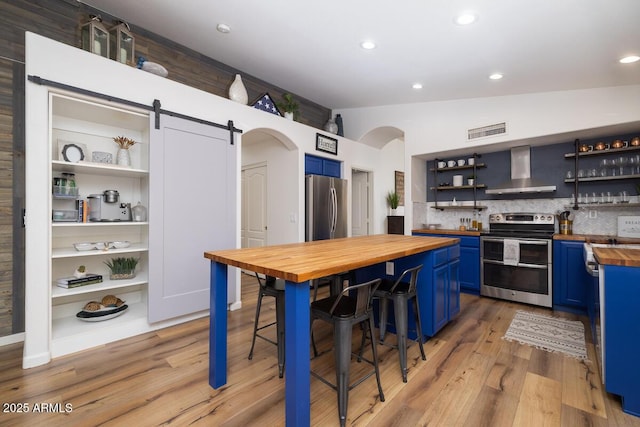 kitchen with blue cabinetry, wall chimney exhaust hood, butcher block countertops, stainless steel appliances, and a barn door