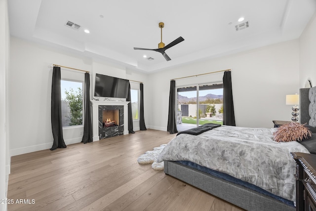 bedroom with a high end fireplace, light hardwood / wood-style floors, access to outside, ceiling fan, and a tray ceiling