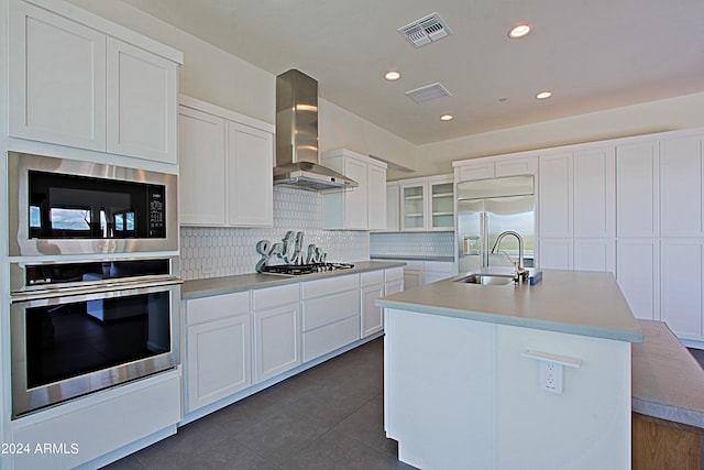 kitchen with wall chimney range hood, an island with sink, sink, built in appliances, and white cabinets