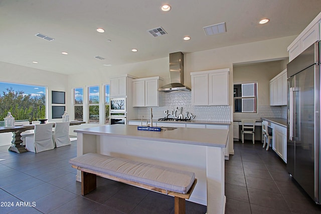 kitchen featuring white cabinets, built in appliances, a center island with sink, and wall chimney range hood