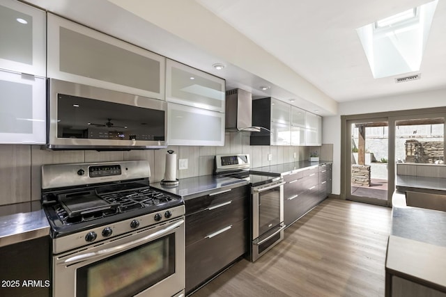 kitchen with stainless steel appliances, stainless steel countertops, backsplash, wall chimney range hood, and modern cabinets