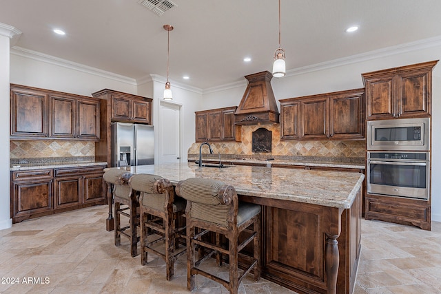 kitchen with stainless steel appliances, custom exhaust hood, an island with sink, crown molding, and decorative light fixtures