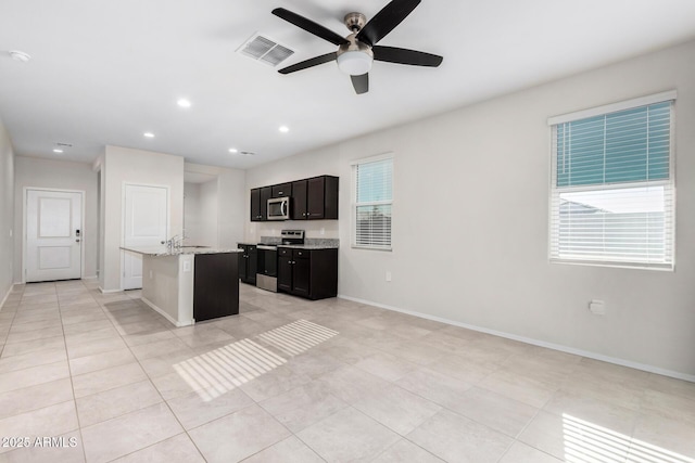 kitchen with light stone countertops, stainless steel appliances, an island with sink, light tile patterned floors, and ceiling fan