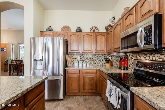 kitchen featuring brown cabinets, light stone counters, arched walkways, appliances with stainless steel finishes, and backsplash