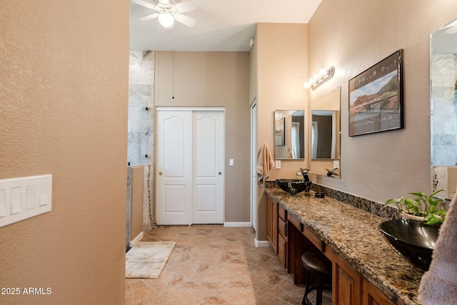 bathroom with double vanity, ceiling fan, a sink, baseboards, and tiled shower
