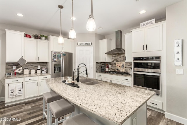 kitchen with sink, an island with sink, wall chimney range hood, and appliances with stainless steel finishes