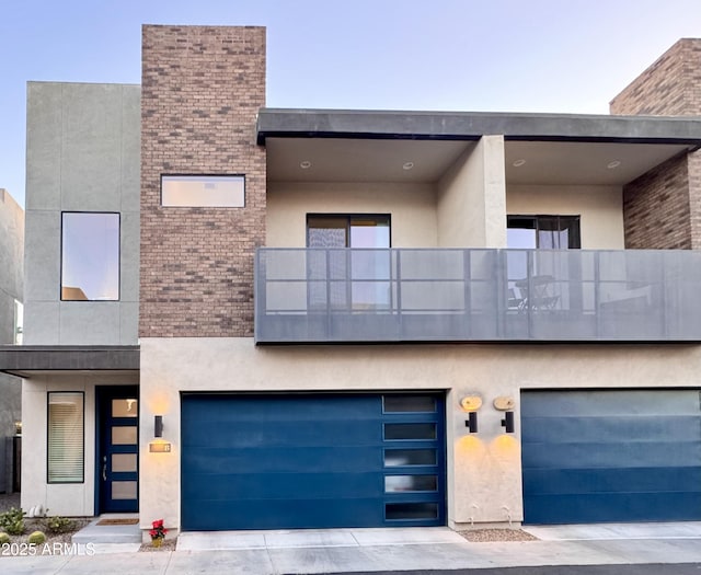 modern home featuring a garage, brick siding, a balcony, and stucco siding