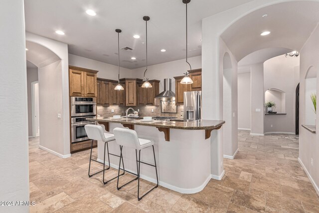 kitchen featuring wall chimney range hood, backsplash, dark stone countertops, pendant lighting, and appliances with stainless steel finishes