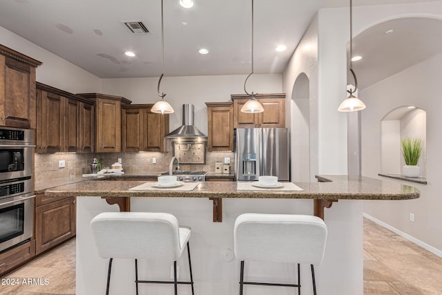 kitchen featuring appliances with stainless steel finishes, hanging light fixtures, wall chimney exhaust hood, and an island with sink