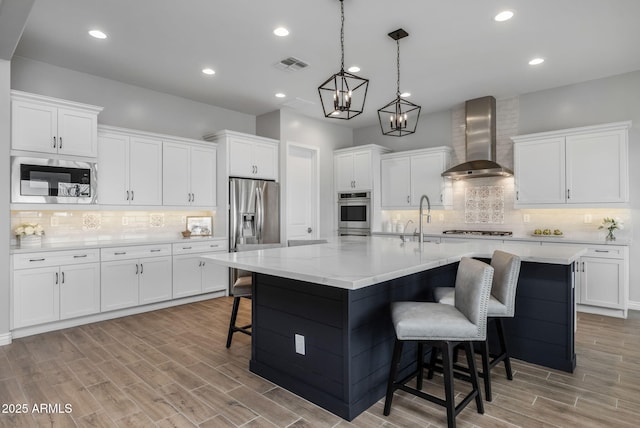 kitchen featuring stainless steel appliances, white cabinetry, a kitchen island with sink, and wall chimney range hood