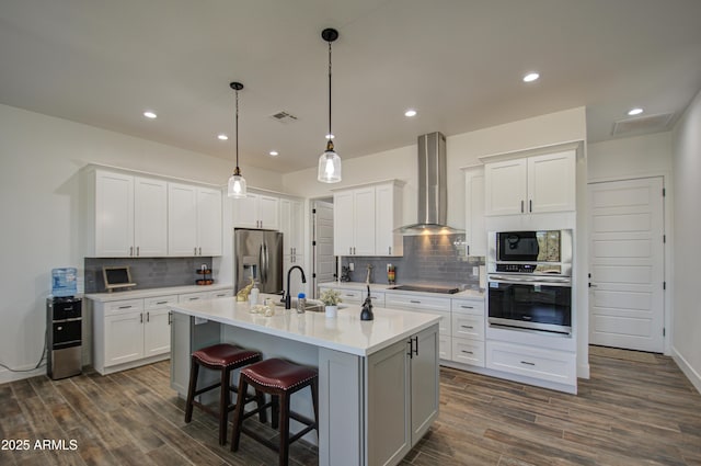 kitchen featuring wall chimney range hood, a kitchen island with sink, white cabinets, stainless steel fridge with ice dispenser, and decorative light fixtures