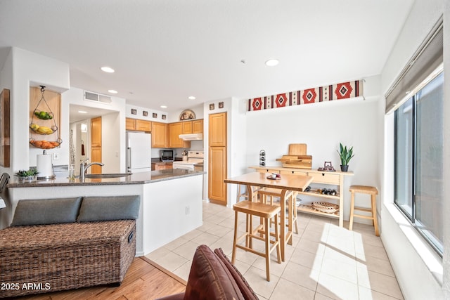 kitchen featuring visible vents, freestanding refrigerator, light brown cabinetry, under cabinet range hood, and electric stove