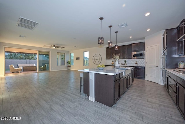 kitchen with stainless steel appliances, hanging light fixtures, dark brown cabinets, and a center island with sink