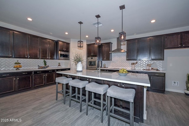 kitchen featuring an island with sink, decorative light fixtures, stainless steel microwave, and wall chimney exhaust hood