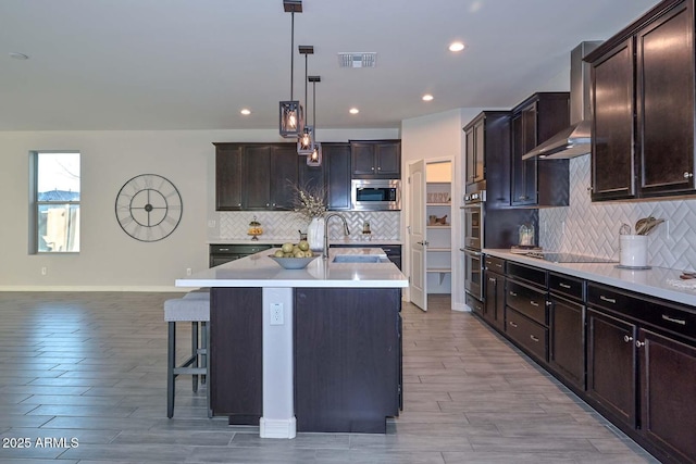 kitchen with sink, appliances with stainless steel finishes, an island with sink, pendant lighting, and wall chimney range hood