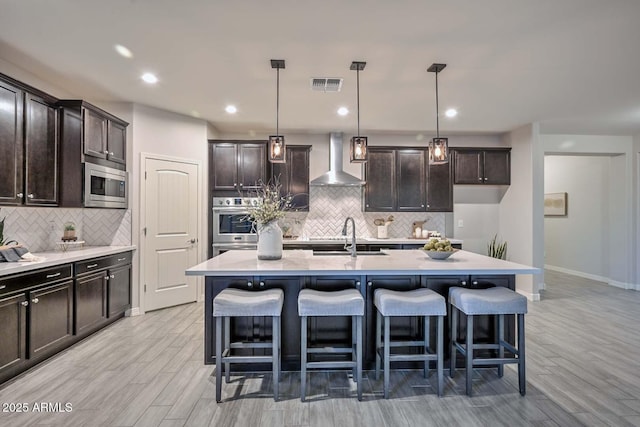 kitchen with pendant lighting, stainless steel microwave, sink, a kitchen island with sink, and wall chimney range hood