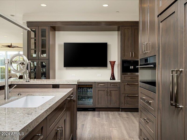 kitchen with ceiling fan, wine cooler, light stone counters, stainless steel oven, and light wood-type flooring