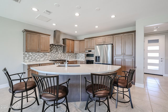 kitchen with stainless steel appliances, wall chimney exhaust hood, sink, a center island with sink, and light tile patterned floors