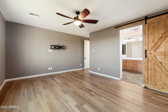 unfurnished bedroom featuring a barn door, light hardwood / wood-style floors, ceiling fan, and connected bathroom