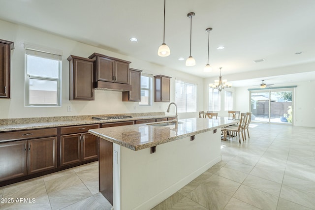 kitchen with a sink, light stone countertops, an island with sink, under cabinet range hood, and stainless steel gas stovetop