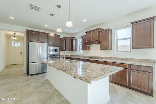 kitchen with appliances with stainless steel finishes, a sink, light stone counters, hanging light fixtures, and an island with sink
