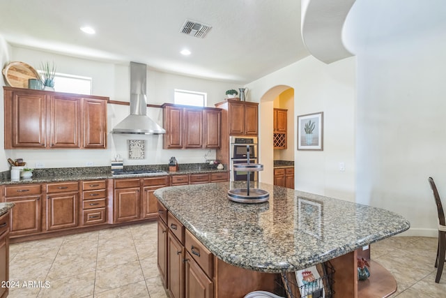 kitchen with wall chimney exhaust hood, dark stone countertops, appliances with stainless steel finishes, and a kitchen island