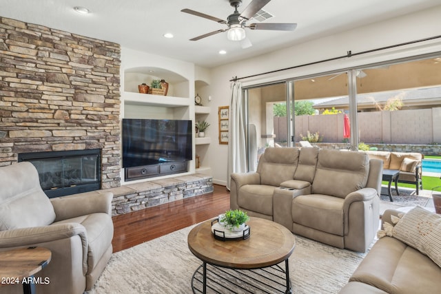 living room featuring built in shelves, a stone fireplace, hardwood / wood-style floors, and ceiling fan