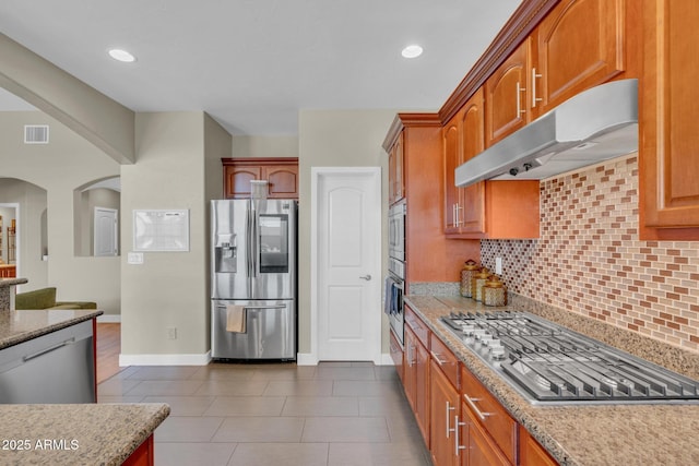 kitchen featuring stainless steel appliances, under cabinet range hood, backsplash, arched walkways, and light stone countertops