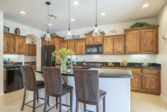 kitchen with brown cabinets, a center island with sink, dark countertops, visible vents, and black appliances