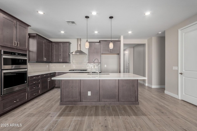 kitchen with double oven, decorative light fixtures, an island with sink, sink, and wall chimney range hood