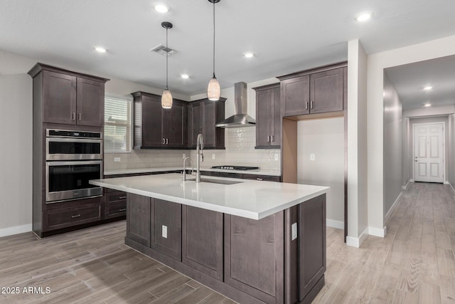 kitchen featuring wall chimney exhaust hood, decorative light fixtures, double oven, black gas cooktop, and a kitchen island with sink