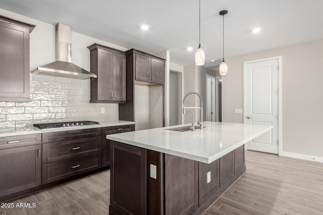 kitchen featuring wall chimney exhaust hood, sink, an island with sink, pendant lighting, and stainless steel gas stovetop